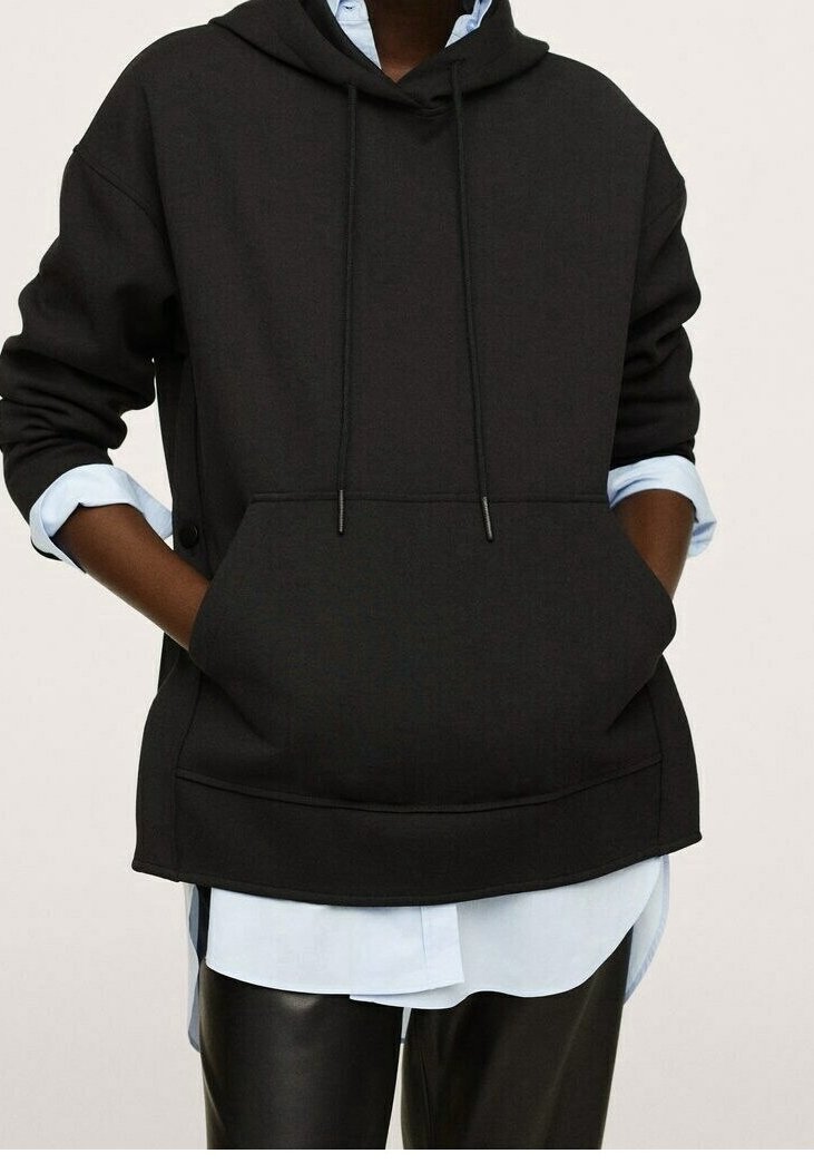 Sudadera negra con capucha, bolsillo frontal, cordones y costuras de hombro caídas, sobre una camisa azul claro y pantalones negros.