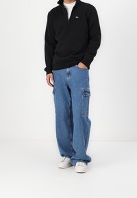Sudadera de forro polar negra con media cremallera, jeans cargo de pierna ancha azules con bolsillos laterales y zapatillas blancas con una parte superior texturizada.