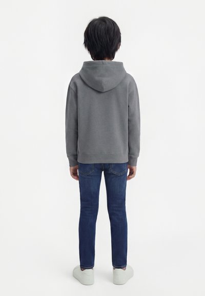 Παιδί που φαίνεται από πίσω, φοράει γκρι φούτερ με κουκούλα, μπλε τζιν και λευκά αθλητικά παπούτσια, στέκεται μπροστά σε απλό ανοιχτό φόντο.