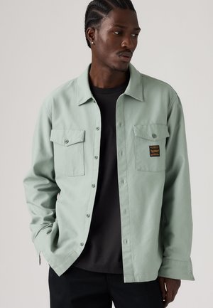 Man met gevlochten haar, die een lichtgroen overhemd met knopen over een zwart T-shirt draagt, kijkt opzij met handen in de zakken.