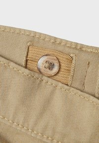 Tissu beige avec des côtes verticales, comprenant une étiquette tissée beige et un bouton rond transparent avec un motif floral. Détails de couture texturée visibles.