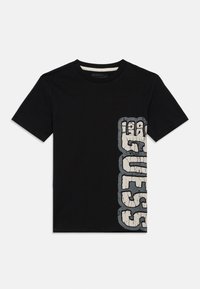 Crna majica kratkih rukava s vertikalno ispucalim bijelim i sivim "GUESS" logotipom s desne strane, okrugli izrez, položena ravno na bijeloj pozadini.