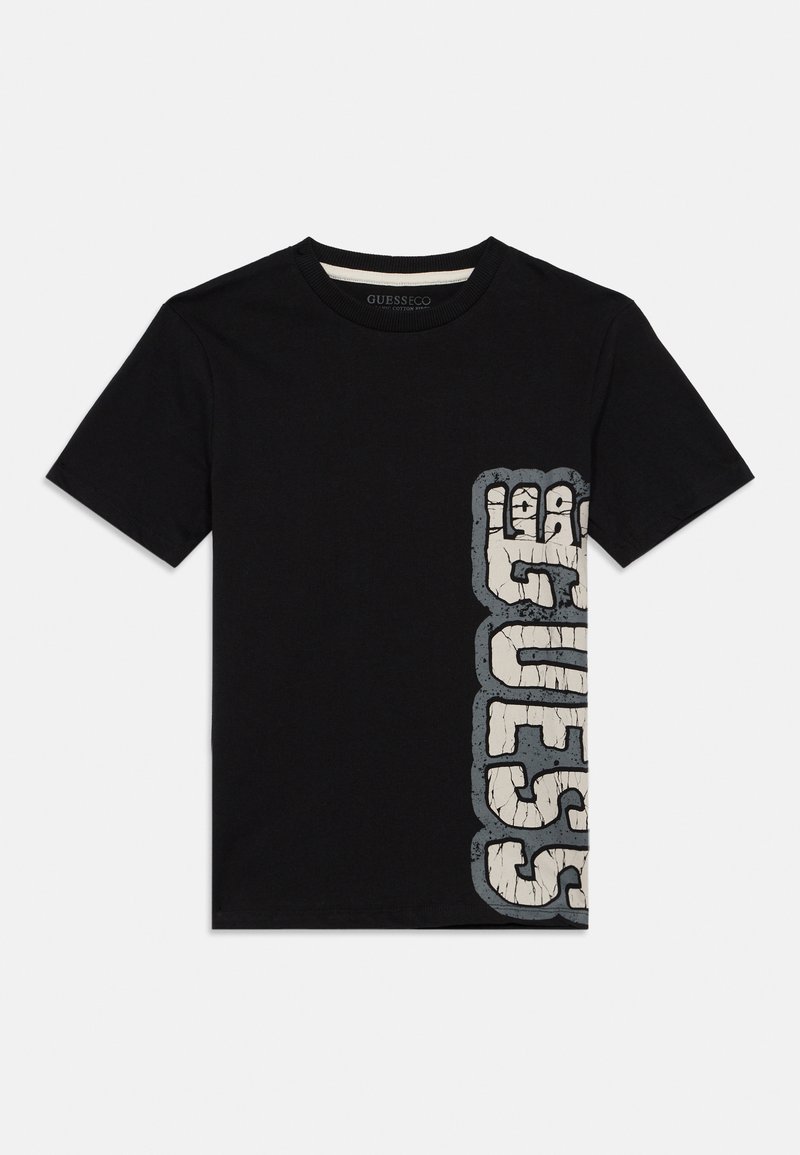 Crna majica kratkih rukava s vertikalno ispucalim bijelim i sivim "GUESS" logotipom s desne strane, okrugli izrez, položena ravno na bijeloj pozadini.