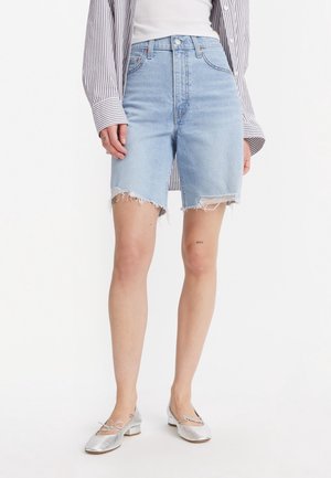 Pantalones cortos de mezclilla azul claro con una cintura alta, dobladillo deshilachado y un ligero desvanecimiento. Combinados con una camiseta sin mangas blanca y una camisa de rayas de gran tamaño. Bailarinas plateadas.