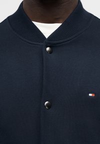 Ζακέτα πλεκτή σε ναυτικό μπλε με ασημί κουμπιά πατιλέτας, ριμπ γιακά, και μικρό λογότυπο σε κόκκινο, άσπρο και μπλε κοντά στο κάτω αριστερό μέρος.