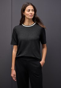 Mujer con cabello largo castaño, vistiendo una camiseta negra de manga corta con un ribete blanco en el cuello y pantalones negros, de pie contra una pared oscura.