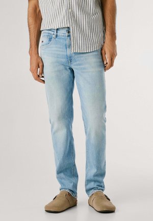 Mand iført lyseblå slim-fit jeans, stribet knappet skjorte og beige slip-on sko, stående mod en ensfarvet baggrund.
