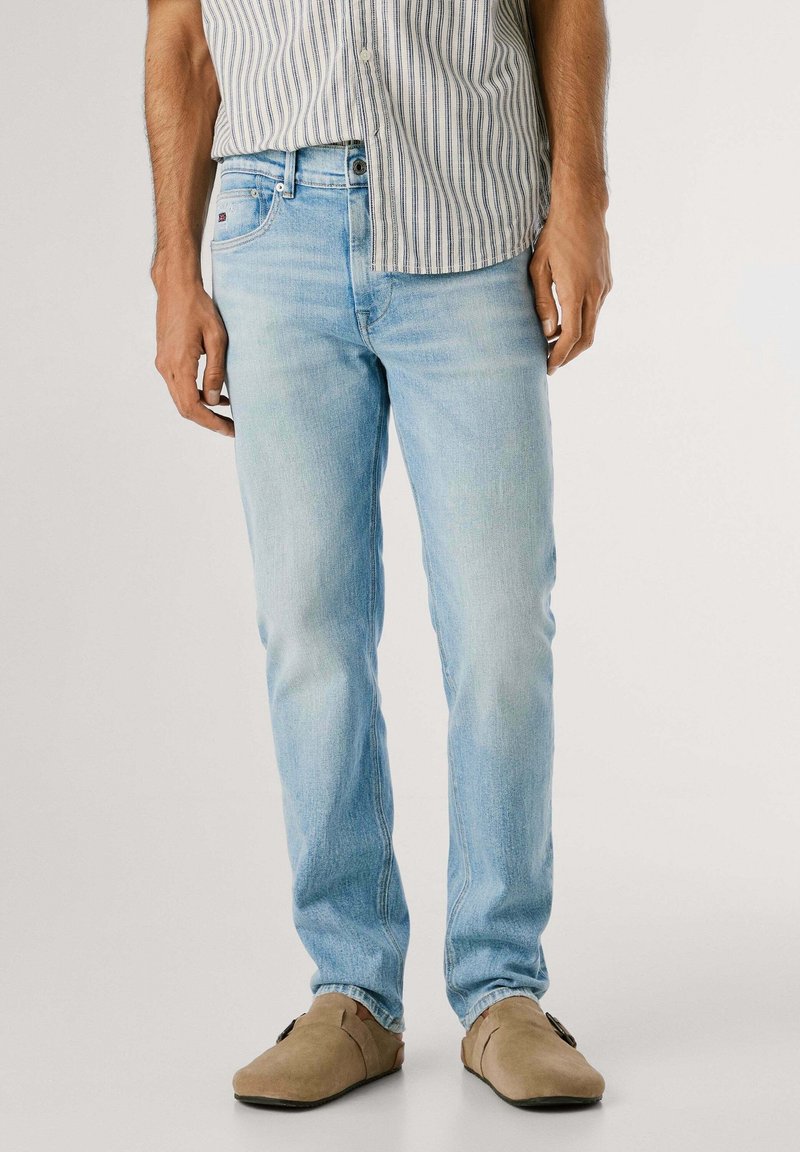 Homme portant un jean slim bleu clair, une chemise à rayures boutonnée et des chaussures beige sans lacets, debout devant un fond uni.