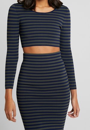 Kvinde iført en langærmet crop top med marineblå og grå vandrette striber og matchende høj-taljet nederdel foran en ensfarvet baggrund.