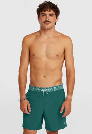 Mężczyzna z wąsami, ubrany w zielone kąpielówki z wzorzystym pasem, stojący na tle jednolitego, jasnego tła.