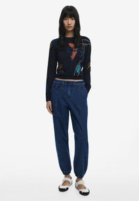 Suéter de punto negro con bordado floral, combinado con pantalones de denim de corte relajado con puños elásticos y bolsillos frontales, complementados con zapatos blancos.