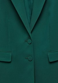 Blazer vert foncé avec deux boutons recouverts de tissu, revers crantés et poches à rabat de chaque côté.