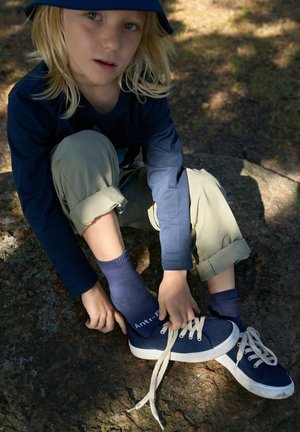 Marineblaue Segelschuhe aus Canvas mit weißen Schnürsenkeln, kombiniert mit marineblauen Socken und hellgrünen Hosen, auf einer felsigen Oberfläche im natürlichen Licht.