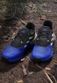 Siniset ja mustat urheilusvajat, joissa on teksturoitu päällinen, paksu kumipohja ja kolme valkoista raitaa sivuilla, luonnollisen maataustalla.