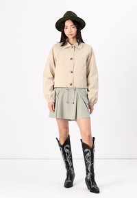 Beige cropped jack met knoopsluiting, lange mouwen en een trekkoordzoom; lichtgroene uitlopende rok; zwarte cowboylaarzen met witte stiksels.