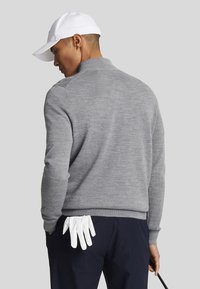 Grau gestrickter Pullover mit hohem Kragen, getragen mit dunklen Hosen. Weiße Golfhandschuhe sind in der Gesäßtasche gesteckt. Das Model ist mit dem Rücken zur Kamera gewandt.