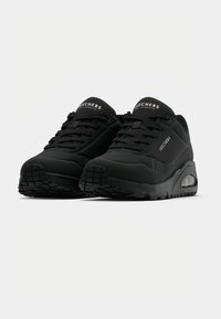 Paire de chaussures de sport noires Skechers avec semelles à coussin d’air visibles et devant à lacets sur fond blanc.