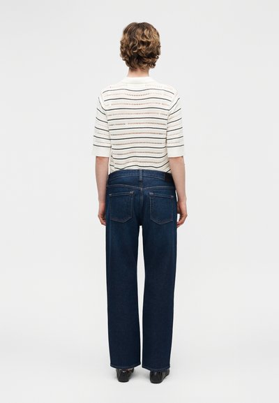 Άτομο με κοντά σγουρά μαλλιά στέκεται γυρισμένο την πλάτη, φορώντας λευκό πλεκτό τοπ με ρίγες και σκουρόχρωμο μπλε τζιν παντελόνι με φαρδιά πόδια καθώς και μαύρα παπούτσια.