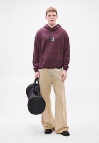 Felpa con cappuccio color mogano con toppa nera con logo, pantaloni beige a gamba larga e scarpe slip-on nere. Tiene una borsa rotonda nera con manici doppi.