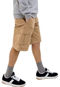 Tannfarbene Cargo-Shorts mit mehreren Taschen, kombiniert mit schwarzen Sneakers mit weißen Akzenten und grauen Socken. Das Modell befindet sich in einer Geh-Pose.