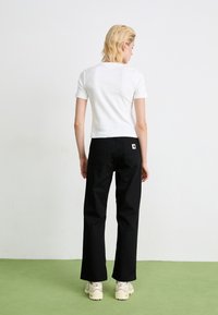 Person with short blonde hair stands on green floor facing white wall, wearing white short-sleeve shirt, black pants, and white sneakers.