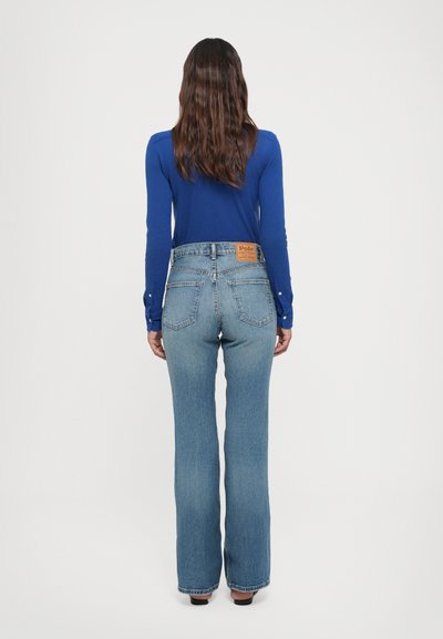 Blå långärmad topp och ljusblå utsvängda jeans, med en midjeetikett och klassisk femfickdesign. Tyget verkar vara denim med lätt slitage.