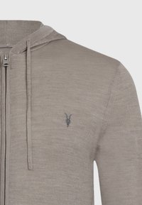 Haut de sweatshirt zippé beige clair en matière douce, arborant un emblème décoratif de crâne de chèvre gris sur la poitrine gauche et des cordons de serrage sur la capuche.