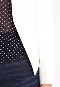 Femme portant un blazer blanc sur un chemisier bleu marine avec de petits motifs d'étoiles blanches, assorti à un jean denim bleu foncé.