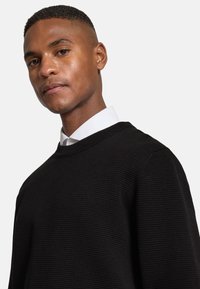 Schwarzer Pullover mit strukturiertem Strickmuster und Rundhalsausschnitt, über einem weißen Hemd mit Kragen getragen. Glatter Stoff mit taillierter Form.