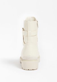 Botines tobilleros de cuero beige con acabado texturizado, con correas laterales y cierre de cremallera. Suela gruesa con la palabra "GUESS" en relieve en el talón.