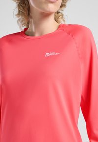 Maglietta atletica a maniche lunghe corallo con collo rotondo. Realizzata in un tessuto liscio e elastico con un piccolo logo bianco sulla parte superiore sinistra.