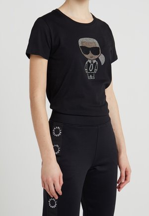 Person iført sort kortærmet skjorte med rhinsten tegneseriefigur i solbriller og sorte bukser med cirkulære "Karl Lagerfeld"-logoer på siderne.