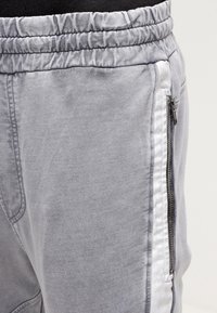 Šedé teplákové kalhoty s páskem na stažení, bočním zipovým kapsou, hladkou texturou a kontrastním bílým bočním pruhem.