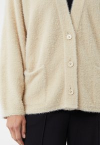 Beżowy puszysty kardigan o fakturowanym wykończeniu, z dekoltem w serek, zapięciem na pięć guzików i dwoma przednimi kieszeniami, noszony na ciemnych spodniach.
