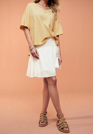 Femme portant un t-shirt jaune ample à col en V, une jupe blanche longueur genou, des sandales beige à fines lanières et des bracelets, debout devant un fond pêche.