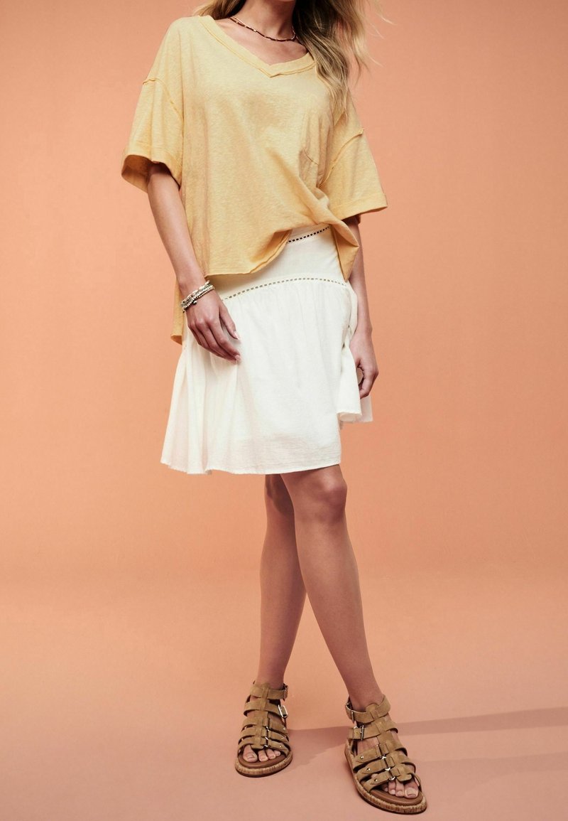 Femme portant un t-shirt jaune ample à col en V, une jupe blanche longueur genou, des sandales beige à fines lanières et des bracelets, debout devant un fond pêche.