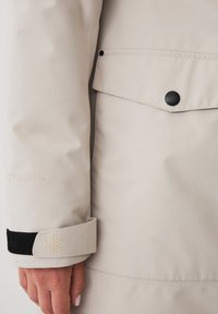 Cappotto beige con una texture liscia, chiusura a pulsante e una tasca. Accenti neri nei polsini con dettagli in rilievo.