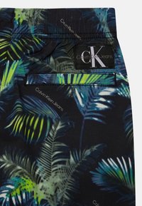 Calças curtas pretas com um padrão de folhas de palma tropicais em verde e azul. Apresenta um bolso traseiro e um rótulo "Calvin Klein Jeans".
