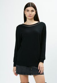 Schwarzer gestrickter Pullover mit weitem Halsauschnitt und Verzierungen entlang des Kragens. Verfügt über lange Ärmel und eine lockere Passform. Kombiniert mit einem Jeansrock.