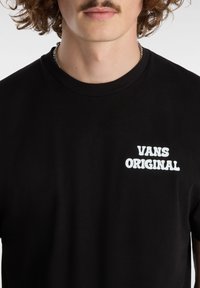 Camiseta de algodón negra con un logo de texto blanco "VANS ORIGINAL" en el área del pecho izquierdo. Manga corta y cuello redondo.