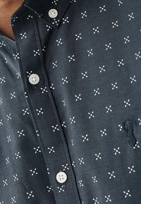 Donkerblauwe shirt met een stoffen patroon van kleine witte bloemachtige ontwerpen en lichtgekleurde knopen op de voorkant.