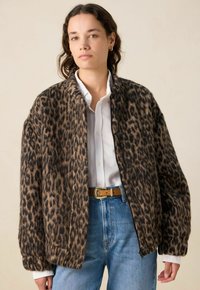 Veste en imprimé léopard en tissu doux, dotée d'une fermeture à boutons, d'une coupe décontractée et de poignets côtelés, portée sur une chemise blanche à boutons.