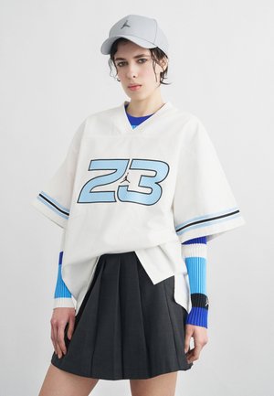 Ung person iført hvid oversized trøje med blåt "23" og Jordan-logo, blåstribede lange ærmer, sort plisseret nederdel og lysegrå kasket.