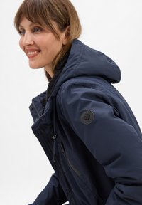 Veste imperméable bleu marine avec capuche, dotée d'une doublure en fausse fourrure noire texturée, de poches latérales et d'un patch logo circulaire sur la manche.