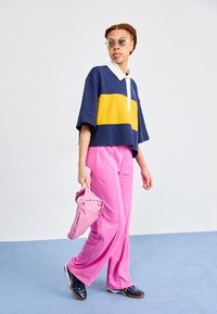 Polo oversized em bloco de cores azul-marinho e amarelo, com gola branca, combinado com calças largas rosas e uma pequena bolsa rosa. Ténis de desporto pretos com detalhes.