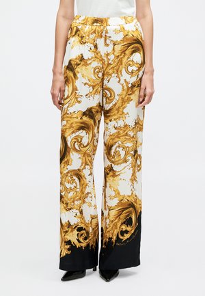 Nő magas derekú, bő szárú nadrágban, amely fehér anyagon sárga és barna barokk mintával van díszítve, fekete szegéllyel, fekete hegyes orrú cipővel párosítva.