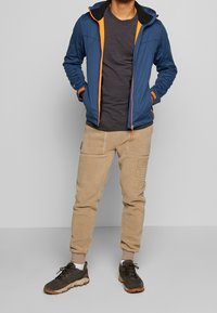 Tenue de plein air pour homme comprenant une veste softshell bleue avec des accents orange, un t-shirt gris, un pantalon en polaire beige et des chaussures de randonnée marron.