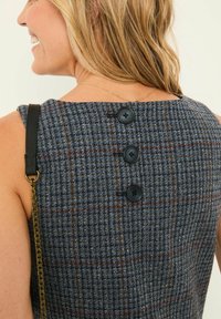 Frau mit blonden Haaren, die ein ärmelloses blaues Karokleid mit drei schwarzen Knöpfen auf dem Rücken trägt und einen schwarzen Taschegurt über der Schulter hat.