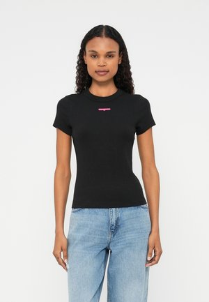 T-shirt nera a righe con maniche corte e scollatura rotonda, con un piccolo logo rosa sul petto. Abbinata a jeans azzurri chiari.