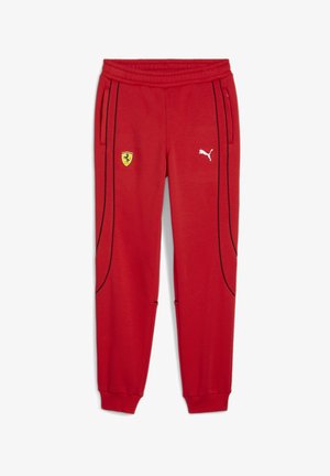 Barn iført rød t-shirt med Ferrari- og Puma-logo, træningsbukser og røde sneakers, stående mod en ensfarvet lysegrå baggrund.